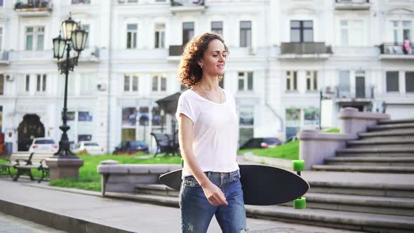 Smiling Attractive Girl Walking in the Old City Street Holding Her Longboard in the Morning Slow alt