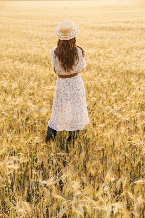 Photo from back of brunette woman in straw hat walking on wheat field
