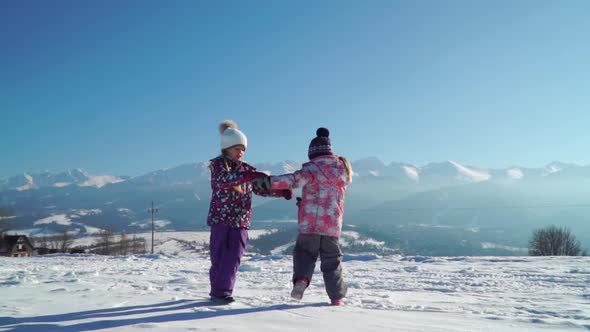 Charming Kids in Outwear Running on Snowy Terrain with Beautiful Mountains on Background in Sunshine