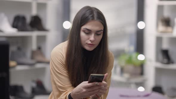 Absorbed Young Caucasian Woman Surfing Internet on Phone Sitting in Shoe Shop Indoors alt