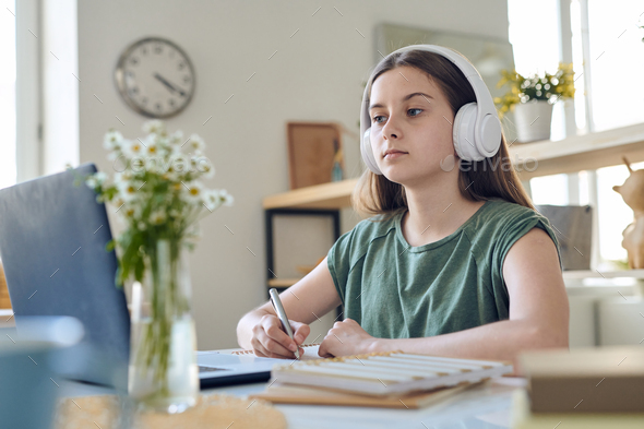 Diligent serious teenage schoolgirl in headphones having remote lesson at home