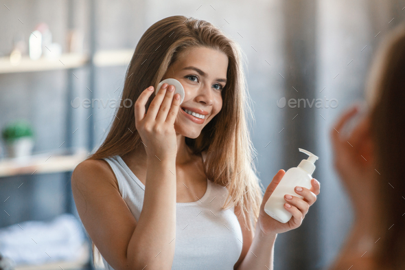 Lovely young woman using makeup remover or facial lotion in front of mirror at bathroom
