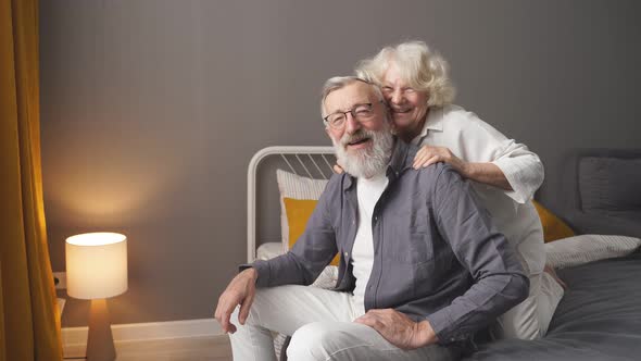Portrait of Happy Retired Couple Sitting at Home in Bedroom Smiling