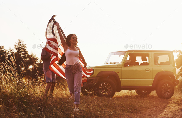 Girl runs forward. Friends have nice weekend outdoors near theirs green car with USA flag