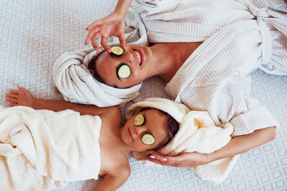 Young positive mother and daughter with towel on head lying on the bed together with cucumber