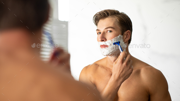 Handsome young man looking in the mirror and shaving