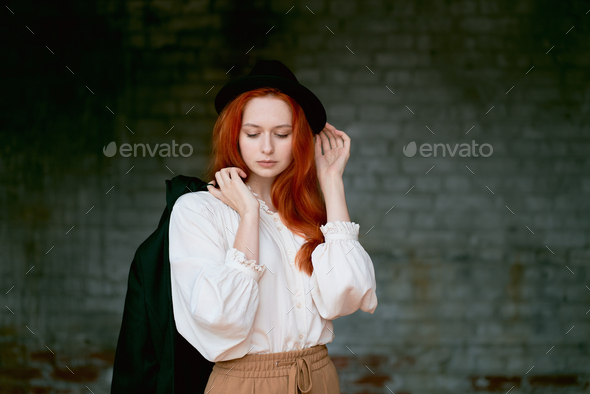 Redhead woman is resting against dark grunge wall. Female in black hat - Stock Photo - Images