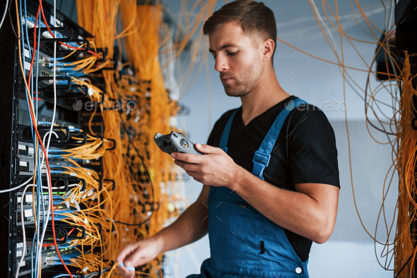 Young man in uniform with measuring device works with internet equipment and wires in server room