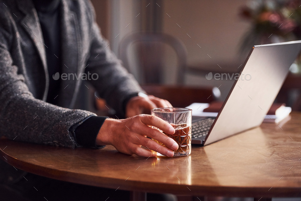 Close up view of elegant young guy in formal wear sits in cafe with his laptop and glass of alcohol