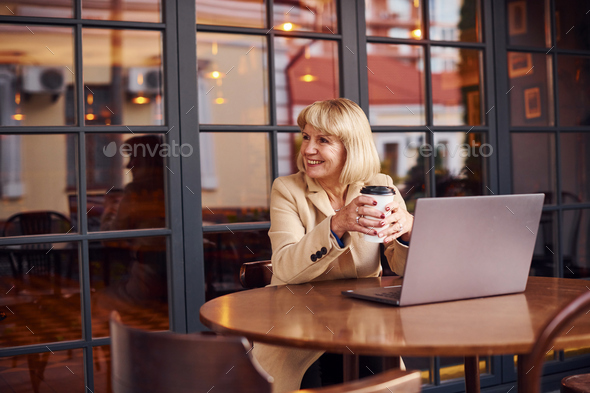 Portrait of senior woman that sits indoors in the cafe with modern laptop