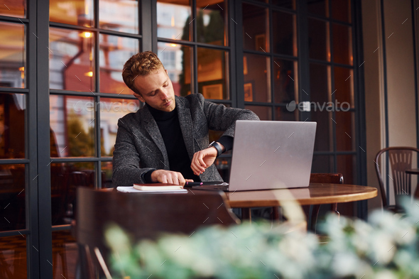 Elegant young guy in formal wear sits in cafe with his laptop and checks time