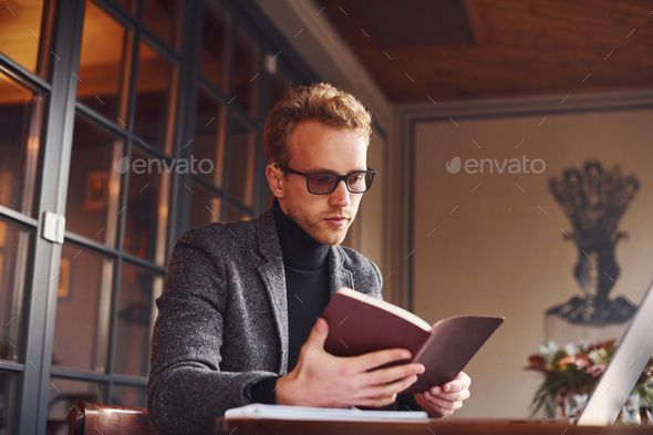 Elegant young guy in formal wear sits in cafe with his laptop and reads the book