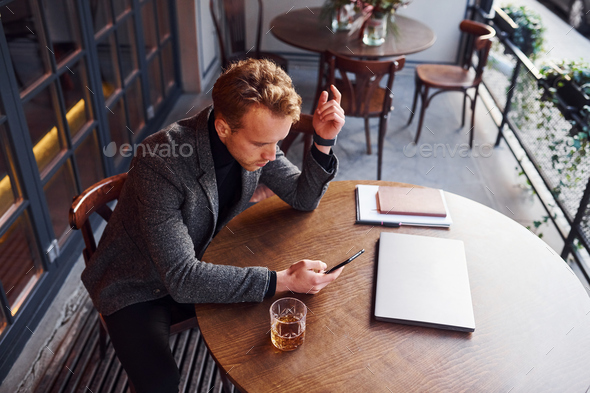 Elegant young guy in formal wear sits in cafe with his laptop and glass of alcohol