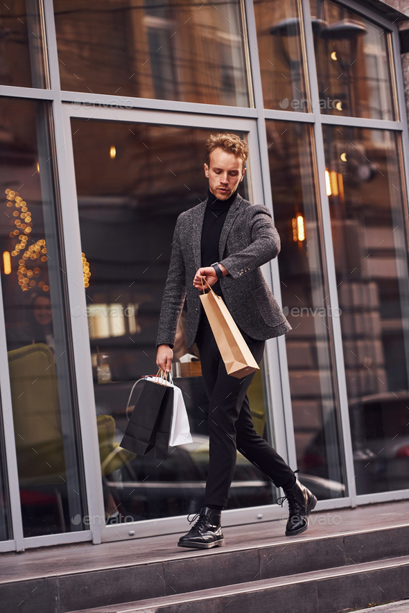 Man in elegant formal wear and with shopping packages is outside against modern building