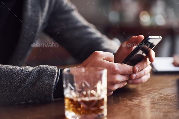 Man in formal wear that sits in the cafe with phone in hands and drink of alcohol on the table