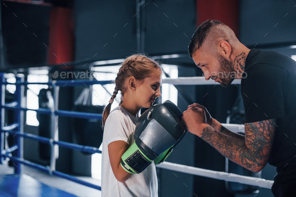 Having sparring on the boxing ring. Young tattooed boxing coach teaches cute little girl in the gym