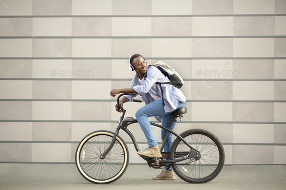 Attractive black guy riding bicycle and listening to music near brick wall outdoors