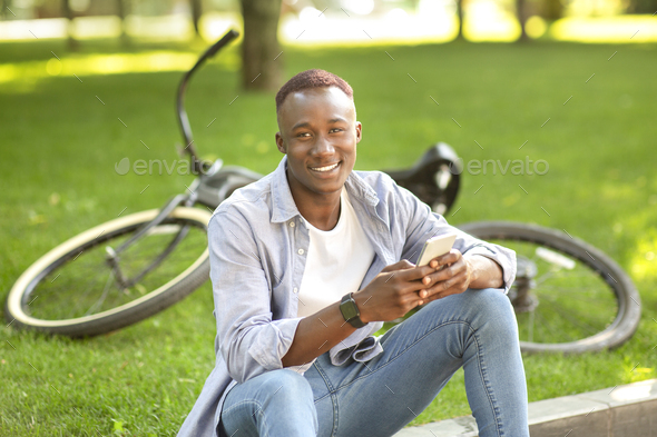 Smiling black guy with mobile phone sitting near his bicycle at park