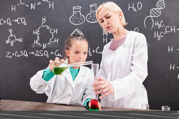 Blond confident teacher of chemistry looking at schoolgirl in whitecoat