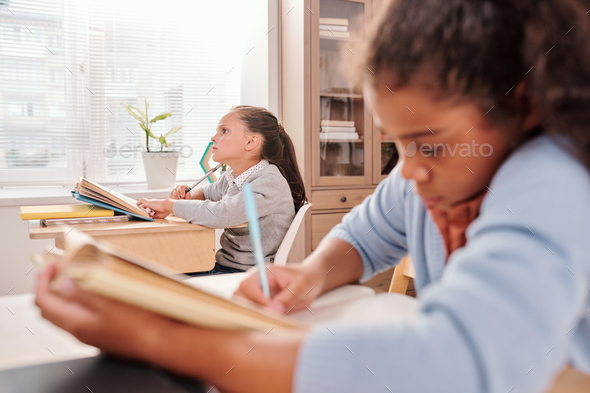 Pensive schoolgirl with open book thinking of answer to question on blackboard