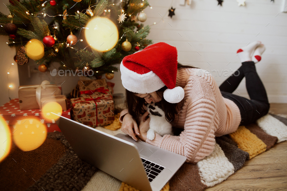 Young woman sitting with cute cat and laptop on rug under christmas tree