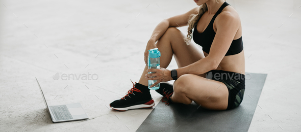 Muscular woman in sportswear with sports tracker sits on mat on floor at home, holds bottle of water