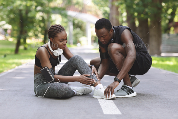 Jogging Injuries. Black Guy Helping Girlfriend Suffering From Sprained Ankle After Running