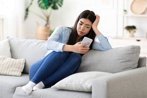 Bored At Home. Weary Asian Girl Sitting With Smartphone On Sofa