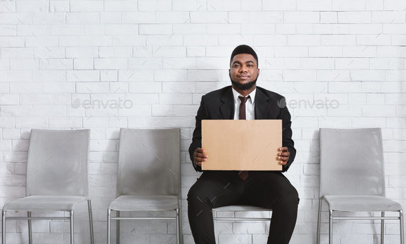 Serious black man in office wear holding blank sign in company hall, waiting for job interview. Copy