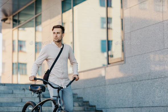 Fast eco city transport. Serious young guy in business clothes, walks with bike at city