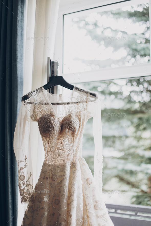 Modern wedding dress hanging at window in soft morning light