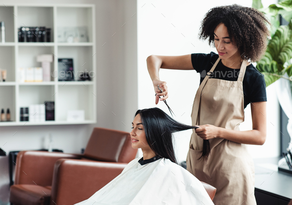 Young black hairdresser cutting hair of female customer at salon