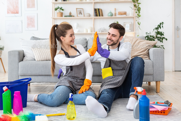 Smiling man and woman in aprons and gloves giving high five in living room with cleaning supplies