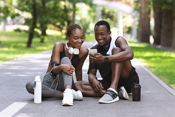 Happy black man and woman relaxing from jogging training outdoors with smartphone