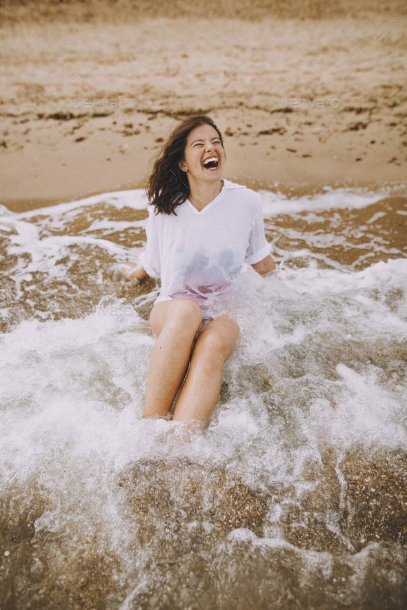 Happy young woman in wet white shirt lying on beach in splashing waves
