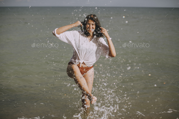 Happy young woman in wet white shirt splashing water drops with leg in sea
