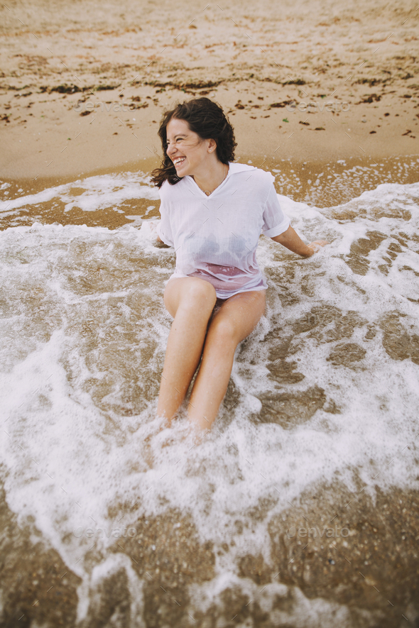 Happy young woman taking off wet white shirt on beach, back view