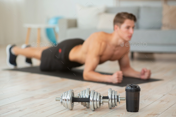 Dumbbells and sports bottle for water close up. Young naked man makes plank on mat