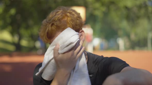 Close-up of Perspiring Young Redhead Sportsman Wiping Face with White Towel Outdoors. Portrait of alt
