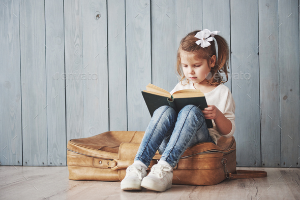 Happy little girl reading interesting book carrying a big briefcase and smiling