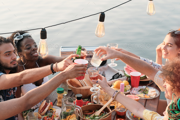 Group of friends sitting at dining table and toasting with cocktails they celebrating the holiday outdoors on a pier