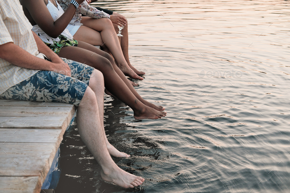 Close-up of group of friends sitting on a pier and wetting their feet in the water in summer day
