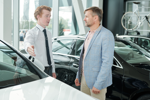 Mature male consumer in casualwear looking at young sales manager pointing at white car and listening to him while choosing new auto
