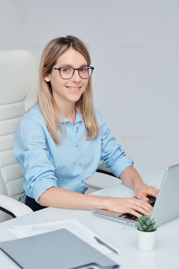 Young successful businesswoman in eyeglasses looking at you with toothy smile while sitting by desk in front of laptop and working in the net