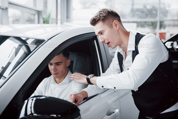 Two men stand in the showroom against cars. Close-up of a sales manager in a suit that sells a car to a customer.