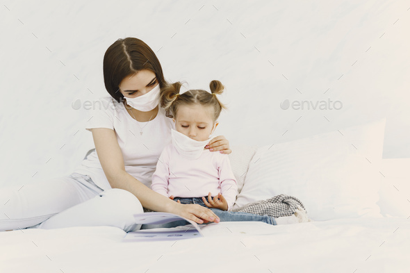 Cute little girl and mother wearing face mask. Family sitting on bed at home. Concept of coronavirus.