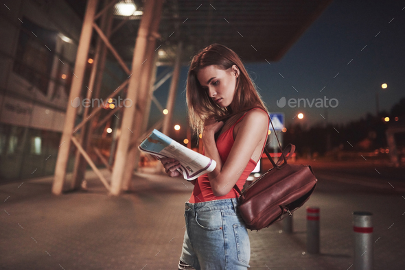 Young girl costs at night near the terminal of the airport or station and reading city map and looking for hotel. Cute tourist with backpacks determine the concept of travel.