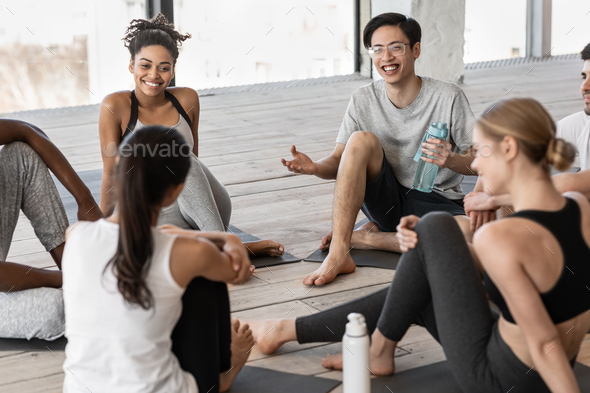 Group of multiracial young people communicating before yoga class in studio, sitting on floor and chatting