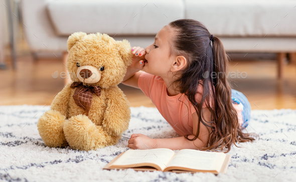 Asian Kid Girl Sharing Secret With Teddy Bear Toy Playing Reading Book Lying On Floor At Home. Best Friend Concept
