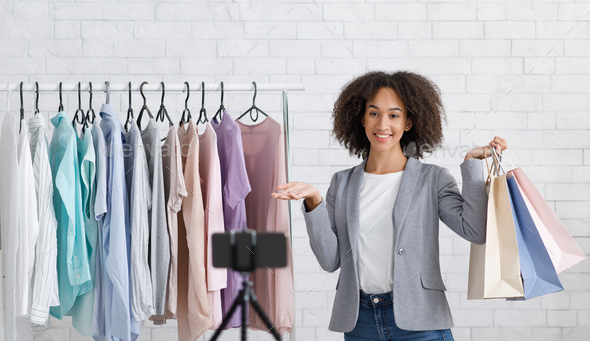 Stylist, personal assistant and hobby online. Smiling african american woman holding packages and shows on clothes rack to web camera in interior, panorama, free space